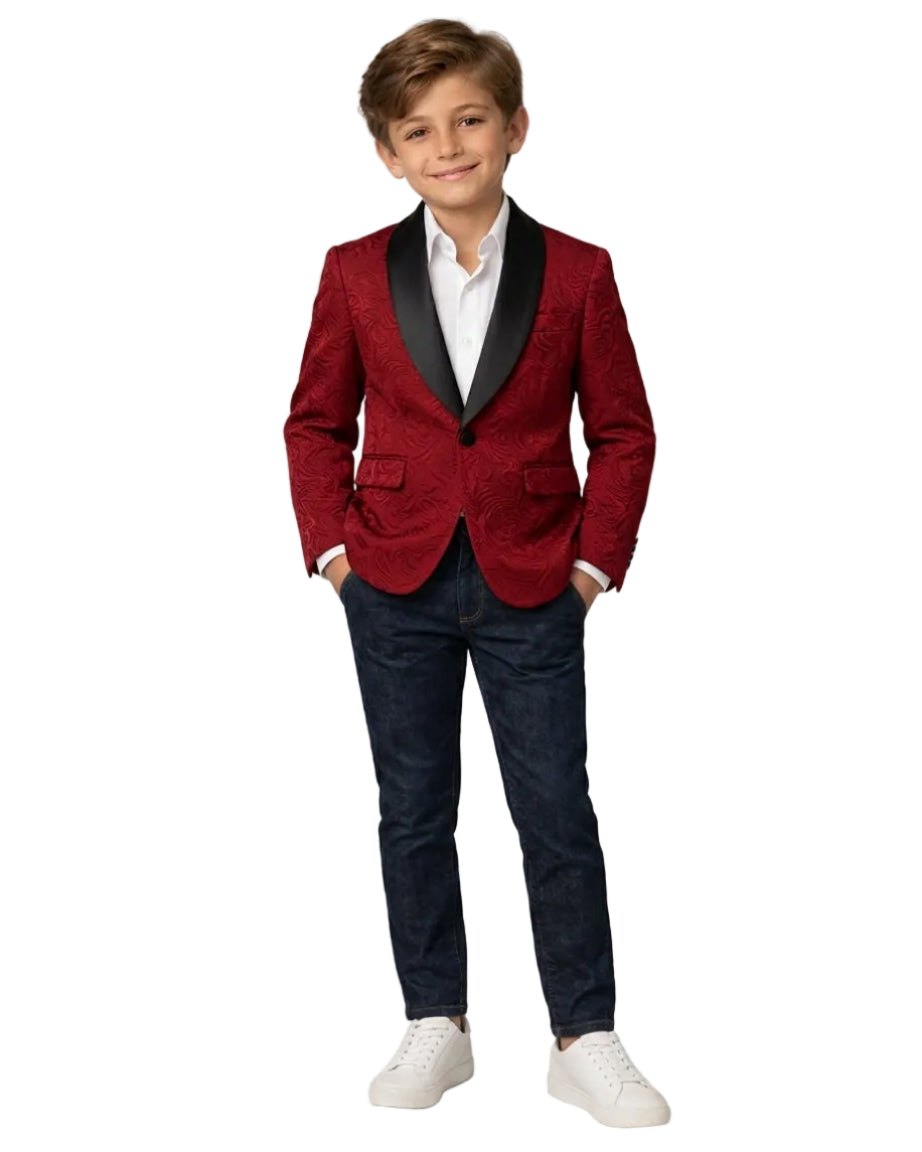 A young boy modeling a red textured blazer with black satin lapels, paired with a white shirt, dark blue jeans, and white sneakers, standing against a light gray studio background.
