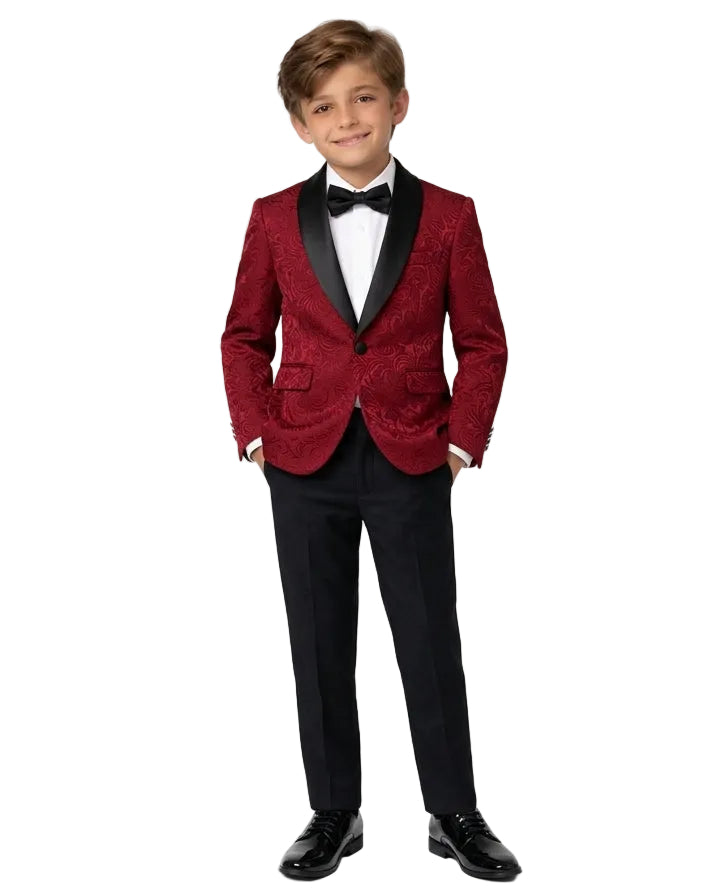 A young boy modeling a formal kids' tuxedo featuring a textured red velvet blazer with black satin lapels, black trousers, a white dress shirt, and a black bow tie, set against a plain white studio background.
