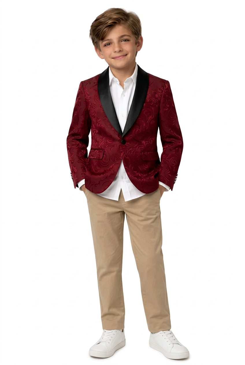 A young boy modeling a red textured velvet blazer with black satin lapels, paired with a white collared shirt, beige trousers, and white sneakers, standing against a plain white studio background.