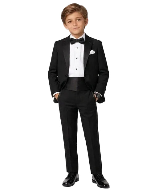 A young boy modeling a black tuxedo with a white dress shirt, black bow tie, and a white pocket square, standing against a plain white studio background.