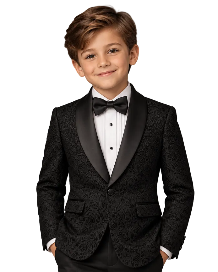 A young boy modeling a black formal tuxedo jacket with a black satin shawl collar, matching black trousers, a white pleated shirt, and a black bow tie, set against a textured gray studio background.