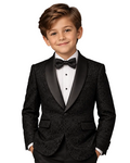 A young boy modeling a black formal tuxedo jacket with a black satin shawl collar, matching black trousers, a white pleated shirt, and a black bow tie, set against a textured gray studio background.