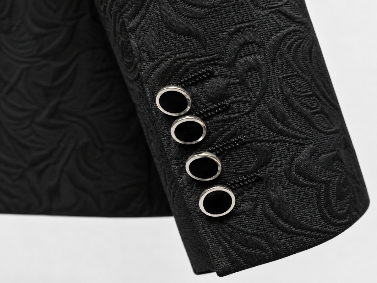 Close-up of a black textured fabric jacket or blazer sleeve with a raised floral jacquard pattern and four silver-tone metal grommets on the cuff, set against a plain white background.