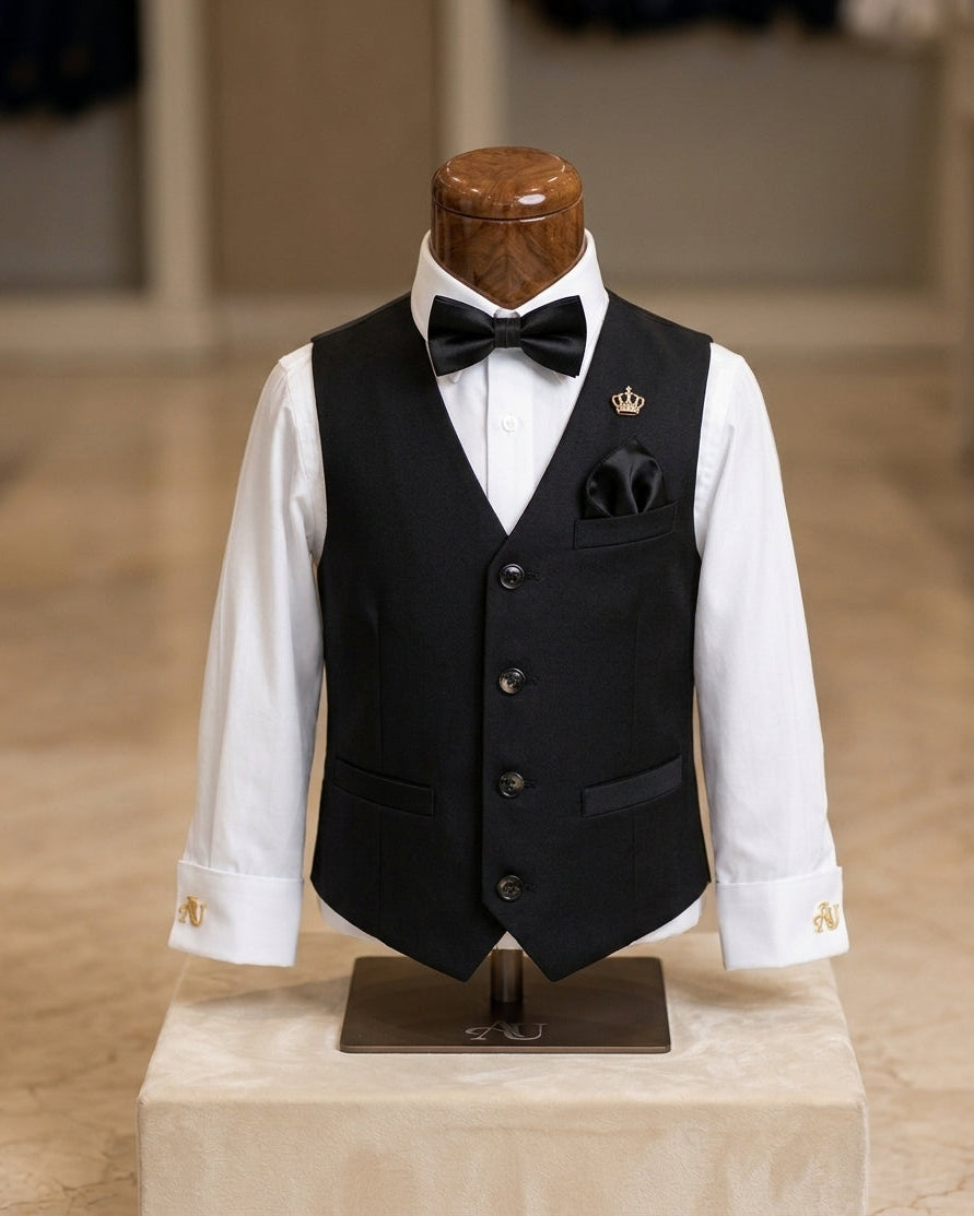 A children's formal outfit displayed on a mannequin torso, featuring a black vest over a white dress shirt with a black bow tie, pocket square, and gold cufflinks. A small gold crown pin is pinned to the chest.
