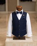 A boy's formal outfit displayed on a mannequin torso, featuring a navy blue vest, matching navy bow tie and pocket square, white dress shirt, and gold cufflinks and lapel pin, set against a softly blurred indoor boutique background.