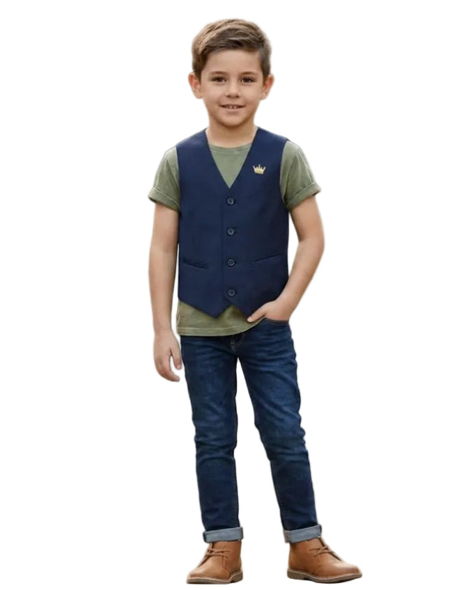 A young boy stands on a stone path in an outdoor playground, wearing a dark navy vest over an olive green t-shirt, blue jeans, and brown boots.